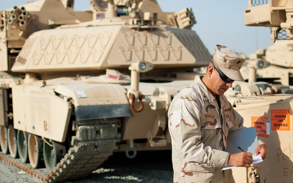 A U.S. military soldier in desert camouflage uniform is writing notes while standing beside a tan-painted armored vehicle, likely during a redeployment or equipment transfer operation. Multiple M1 Abrams tanks are visible in the background, parked on a gravel surface in what appears to be a staging or logistics area. Text written on the vehicle reads 'FT. HOOD', suggesting equipment is being processed for return to Fort Hood, Texas.