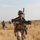 A U.S. Marine in full combat gear walks through a dry, grassy field toward the camera, carrying an M16/M4-style rifle. Two additional armed personnel are visible in the background, suggesting a patrol formation. The terrain and uniforms are consistent with a desert or arid operational environment.