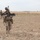 The image shows military personnel conducting a foot patrol across an arid, open terrain. The lead soldier is prominently visible in the foreground carrying a rifle in a ready position, with at least two additional soldiers visible in the background. The environment appears to be a dry, flat desert or semi-arid landscape consistent with operational deployment conditions.