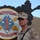 A U.S. Marine in desert MARPAT (MCCUU) uniform stands in front of a unit insignia sign for 2nd Battalion, 11th Marines, 1st Marine Division. The sign displays the unit motto 'Second to None' and battle honor 'Guadalcanal,' mounted on a black painted wooden board. Text below the emblem references 'Firebase' and partially visible location text, suggesting a forward operating base setting.