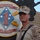 A young male service member in USMC MARPAT desert camouflage uniform stands in front of a unit insignia plaque. The plaque belongs to 2nd Battalion, 11th Marines, 1st Marine Division, bearing the motto 'Second to None' and the battle honor 'Guadalcanal'. The image appears to be taken at a military installation, likely in a deployed or forward operating environment.