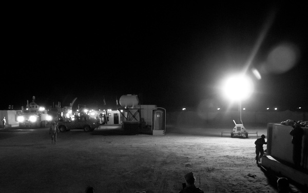 A black-and-white nighttime photograph of what appears to be a military forward operating base or staging area. Multiple personnel in military-style gear are visible in the foreground and background, with vehicles, temporary structures, and a bright artificial light source illuminating the scene. The setting appears to be an arid, open terrain compound.