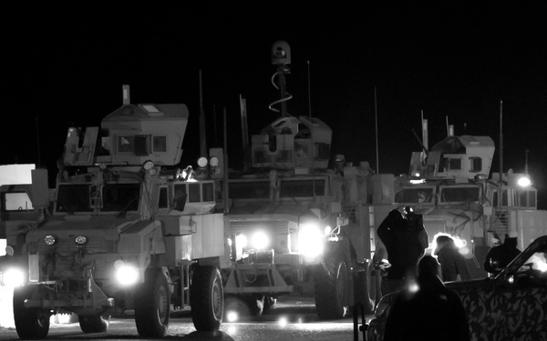 A nighttime black-and-white photograph showing a convoy or staging area with multiple military armored vehicles, likely MRAPs or similar mine-resistant ambush-protected vehicles. Several personnel are visible on foot moving around the vehicles, which have their headlights illuminated. A camouflage-patterned vehicle is visible on the right side of the frame.