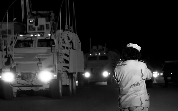 A nighttime military scene showing a soldier in desert camouflage uniform standing on a road, facing an approaching convoy of military vehicles. The soldier appears to be directing or monitoring the convoy's movement. The image is captured in black and white, with vehicle headlights providing the primary illumination.