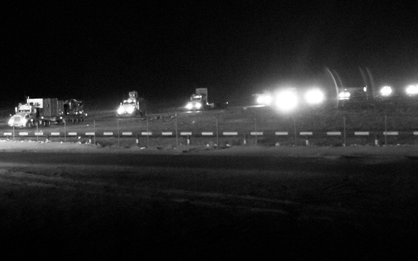 A nighttime black-and-white photograph showing a military or industrial convoy of vehicles lined along a road or perimeter. Multiple vehicles with headlights and work lights are visible stretching across the middle distance. The foreground contains coiled concertina/razor wire, suggesting a secured or fortified position.