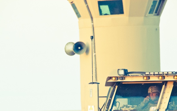The image shows a large tan/desert-colored military armored vehicle, likely an MRAP (Mine-Resistant Ambush Protected) vehicle, in what appears to be a forward operating base or checkpoint environment. A soldier wearing a helmet is visible through the windshield of the vehicle. The setting appears to be an arid, desert region based on the terrain and color palette.