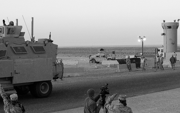A black and white photograph showing military personnel waving at a large armored vehicle, likely an MRAP (Mine-Resistant Ambush Protected vehicle), at what appears to be a military base checkpoint or gate. Several soldiers and a camera operator are visible in the foreground, while guard towers and additional military vehicles are visible in the background. The scene suggests a convoy departure or arrival being documented.