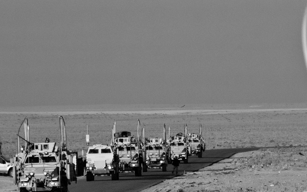 A black-and-white photograph showing a military convoy of armored vehicles traveling along a paved road through an arid desert environment. The convoy consists of multiple MRAPs or similar mine-resistant vehicles equipped with antennas and communications equipment. The image appears to be taken from behind a perimeter security barrier, as concertina/razor wire is visible in the foreground.