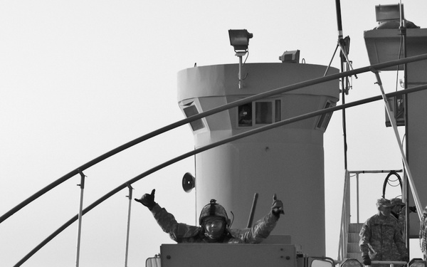 A black and white photograph depicting military personnel in what appears to be a forward operating base or checkpoint environment. A soldier is visible in the turret of an armored vehicle, gesturing with both hands raised. Additional personnel in military gear are visible in the surrounding area.