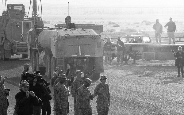 A black and white photograph depicting a military gathering or ceremony in what appears to be a desert environment. Multiple soldiers in camouflage uniforms are assembled, with several individuals appearing to shake hands or interact formally while media personnel with cameras document the event. Large military vehicles and trucks are visible in the background.