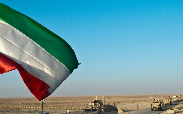A military convoy of tan/desert-camouflage armored vehicles is passing through a checkpoint in an arid desert environment. The Kuwaiti flag is prominently displayed in the foreground, flying from a flagpole surrounded by razor wire. The scene depicts a border crossing or military entry/exit point with concrete barriers and yellow warning tape marking the perimeter.