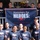 'Hammers for Heroes' gets hand from Marines and sailors A group of approximately 18-20 volunteers pose together holding a banner reading 'Hammers for HEROES: Remembering Pearl Harbor' in front of what appears to be a residential building under renovation. The participants are wearing matching navy blue 'Hammers for Heroes' t-shirts, suggesting an organized community service or charity build event. The group includes men and women of various ages, with some wearing construction hard hats, indicating active work on the property.