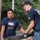 'Hammers for Heroes' gets hand from Marines and sailors Two young men are working together outdoors in rainy conditions, lifting or moving a heavy piece of debris or material that appears to be old asphalt or concrete. The man on the left wears a 'Hammers for Heroes: Remembering Pearl Harbor' t-shirt featuring Habitat for Humanity and Lowe's logos, indicating this is a volunteer community service event. The man on the right wears safety glasses, a baseball cap, and Mechanix brand work gloves.