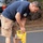 'Hammers for Heroes' gets hand from Marines and sailors A young man is bent over operating a yellow DeWalt rotary hammer drill into the edge of an asphalt road surface. He is wearing a navy blue t-shirt, khaki cargo shorts, black boots, and sunglasses pushed up on his head. A vehicle is partially visible in the background along with a rocky embankment.