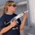 'Hammers for Heroes' gets hand from Marines and sailors A young woman wearing a navy blue t-shirt and sunglasses is holding a DAP caulk gun, appearing to inspect or prepare to apply caulking to a building exterior. Her shirt displays logos for Habitat for Humanity and Lowe's Home Improvement, suggesting she is participating in a volunteer construction or home repair project. She is standing near the exterior wall and window trim of a gray-painted structure.