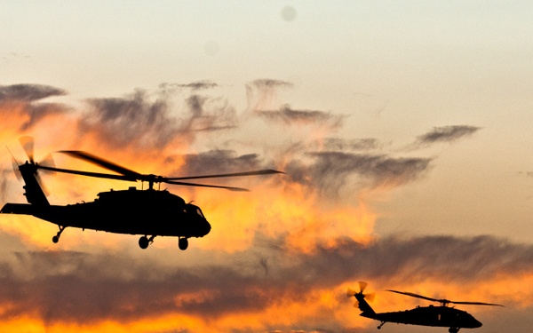Two military helicopters are captured in silhouette against a dramatic sunset sky with vivid orange and golden clouds. The helicopters appear to be UH-60 Black Hawks flying in loose formation. The backlighting from the sunset creates a striking contrast between the dark aircraft silhouettes and the illuminated sky.