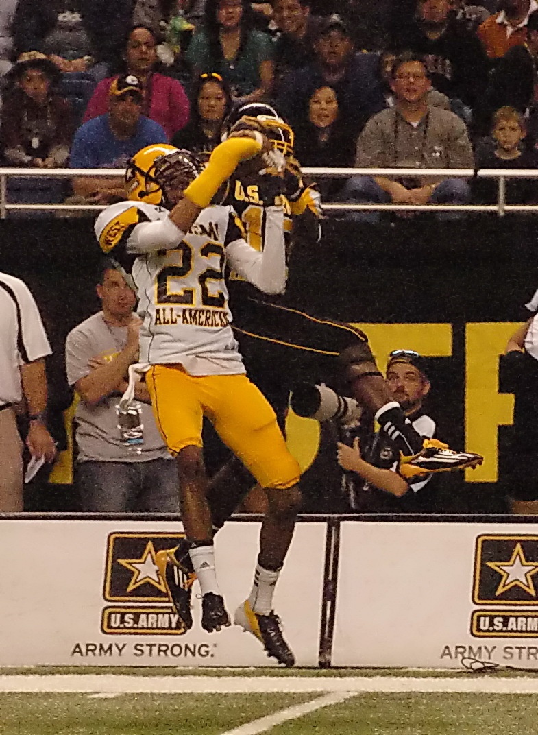 A football game action shot showing two players competing for a pass near the end zone. One player wearing a white 'All-American' jersey numbered 22 with gold pants is contesting a catch against a player in a dark uniform numbered 11. The game appears to be the U.S. Army All-American Bowl, as indicated by the branding on the field barrier.