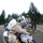 Military personnel in CBRN (Chemical, Biological, Radiological, Nuclear) protective gear are conducting a decontamination training exercise. A simulated casualty lies on a stretcher while personnel in MOPP suits and gas masks perform decontamination procedures using a sprayer backpack unit. The scene takes place on a gravel surface with conifer trees in the background, suggesting a Pacific Northwest training environment.