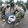 Military personnel in CBRN (Chemical, Biological, Radiological, Nuclear) protective gear and gas masks are conducting a casualty decontamination training exercise. A simulated casualty in civilian clothing is lying on a military litter/stretcher on a gravel surface. Multiple soldiers in woodland camouflage BDUs and tan MOPP suits are working together to manage the mock victim.