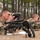Two soldiers in MultiCam/OCP camouflage uniforms are lying prone on the ground, looking through optical observation devices, with scoped rifles positioned in front of them on bipods. A third soldier in ACU (Army Combat Uniform) stands in the background appearing to supervise or evaluate. The setting appears to be a wooded training range, likely a sniper or reconnaissance training exercise.
