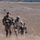 A group of soldiers in full combat gear are moving across an open, arid training field. The foreground shows two primary soldiers carrying rifles and wearing tactical vests and helmets, with additional soldiers visible in the background. The setting appears to be a military training exercise on a large open range.
