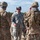 A group of U.S. Army soldiers are gathered outdoors at what appears to be a training area or military installation. A central figure wearing Army Combat Uniform (ACU) in Universal Camouflage Pattern (UCP) is engaged in conversation with soldiers wearing Operational Camouflage Pattern (OCP/Multicam) uniforms and full combat gear. The setting suggests a field training exercise or briefing.
