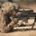 A U.S. Army soldier identified as PFC Howerton is in a prone firing position aiming a semi-automatic rifle equipped with a telescopic scope and bipod. The soldier is wearing MultiCam (OCP) camouflage uniform and tactical gloves on what appears to be a desert training range. Additional soldiers are visible in the blurred background in similar positions.