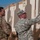 Two military personnel in camouflage uniforms are examining shooting range targets made of cardboard. One soldier wearing a patrol cap and glasses is marking or inspecting target number 8 with a pen or marker, while the other soldier wearing a backpack observes. The setting appears to be an outdoor military firing range under a partly cloudy sky.