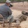 The image shows a military training or qualification range where soldiers in camouflage uniforms are in prone firing positions with rifles equipped with bipods and optics. A person wearing a gray sweatshirt and military camouflage pants, with a name tag visible on their cap, is crouching nearby using a spotting scope or camera with a telephoto lens. The setting appears to be an outdoor range with sandy ground and sparse trees in the background.