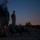 A military training exercise photographed at dusk or dawn showing a prone shooter in the foreground aiming a rifle equipped with an optical sight. Several standing personnel in military uniforms observe from the background near a structure with visible light. The low-light conditions and overall scene suggest a night or twilight live-fire training exercise.