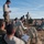 A group of military personnel are gathered in an open field conducting what appears to be a briefing or after-action review. The soldiers are wearing a mix of camouflage uniforms and tan/beige base layers, with some seated on the ground and one standing. Several individuals are taking notes on notepads or referencing handheld devices.