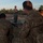 A military instructor in a boonie hat and camouflage uniform addresses a group of soldiers seated on the ground during an outdoor field training session. The soldiers, wearing MultiCam/OCP pattern uniforms, appear to be attending a briefing or instruction period, with at least one taking notes on a notepad. The setting appears to be a training range or open field area during early morning or late afternoon light.
