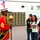 A U.S. Marine in full dress uniform is interacting with a group of students in what appears to be a school classroom or band room. The Marine is holding a ceremonial mace and wearing a distinctive red dress uniform with a white cover (hat) adorned with a small American flag. The students appear engaged and attentive during what seems to be a school visit or educational presentation.