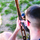 An overhead view of a young male with a short buzz cut holding a compound bow, viewed from above and behind. He appears to be aiming or preparing to shoot the bow outdoors on a sandy, patchy grass surface. The image captures close detail of both the bow's riser and the person's grip.