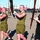 A group of shaved-head military personnel, likely U.S. Marine Corps recruits or drill team members, are practicing rifle drill movements in a paved outdoor area. Three individuals are prominently visible in the foreground, wearing olive green or black athletic attire and handling rifles with bayonets attached. The setting appears to be a military base or training facility, with vehicles, fencing, and structures visible in the background.
