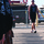 The image shows a group of individuals wearing matching black 'United States Marine Corps Silent Drill Platoon' shirts holding rifles in a line formation. A single person in a matching black tank top and shorts, wearing sunglasses, walks toward the formation. The scene appears to take place outdoors on a paved surface near a chain-link fence.