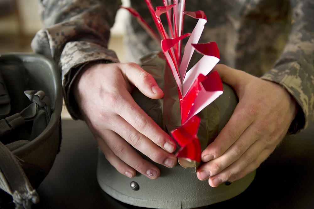 Two individuals wearing US military camouflage uniforms (ABU/ACU pattern) are handling a green cylindrical object with red and white safety streamers or flags attached to the top. The red streamers are characteristic of safety/training markers used on military ordnance or training devices to indicate inert or safe status. A military helmet is visible in the background to the left.