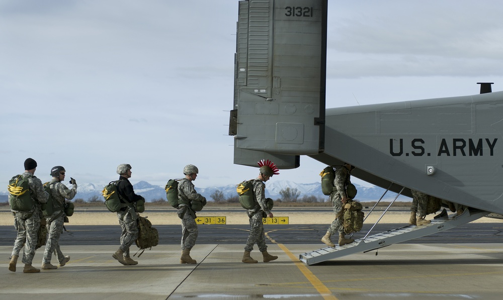 A group of U.S. Army soldiers in combat uniforms and full gear are boarding a military aircraft via a rear loading ramp. The soldiers are carrying large green rucksacks and personal equipment. One soldier is notably wearing a red mohawk-style headdress, likely a traditional pre-deployment or airborne tradition.