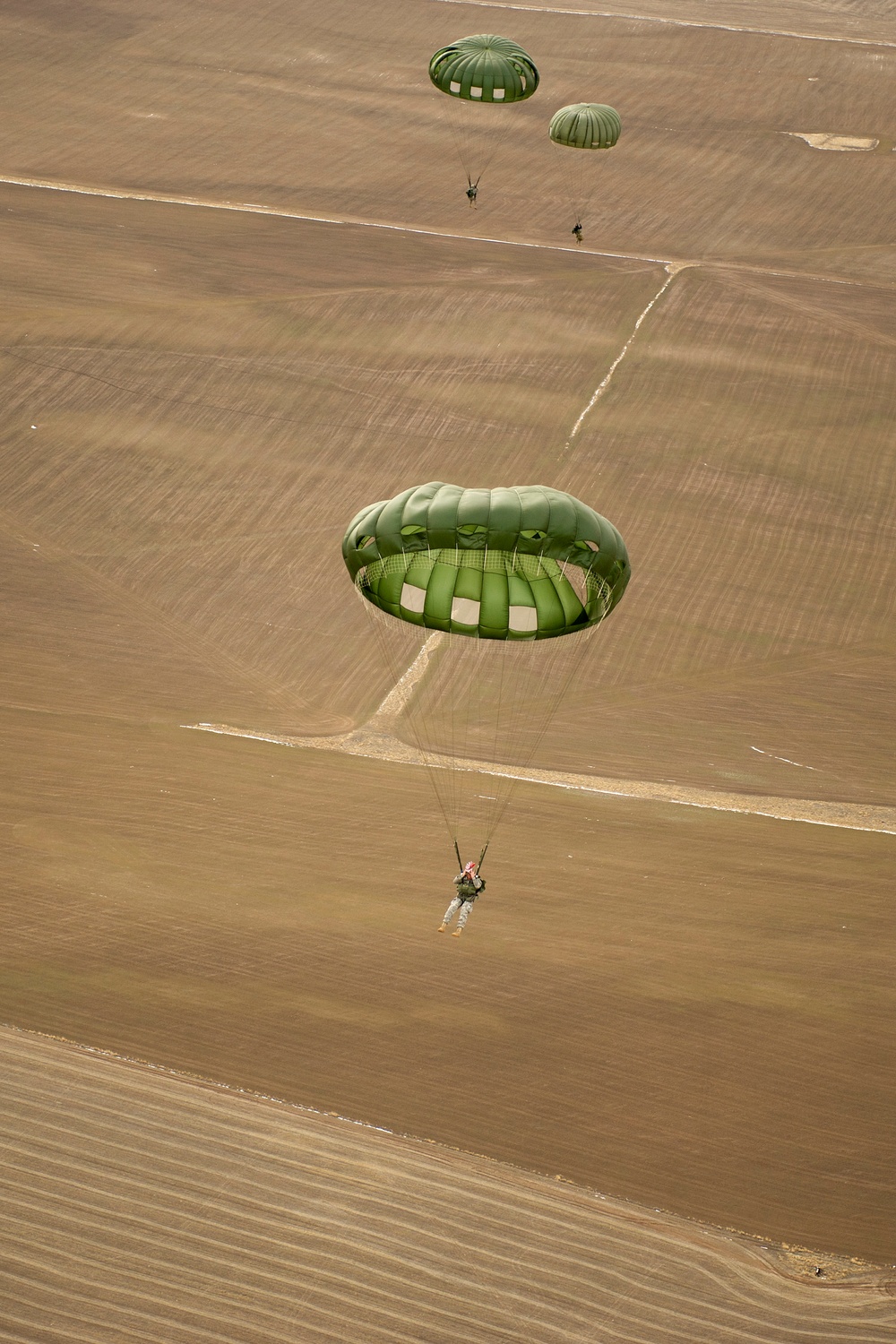 An aerial photograph captures multiple military paratroopers descending over a large, flat agricultural field with visible tractor furrow patterns. At least three green military parachutes are visible at different altitudes, suggesting a sequential or mass tactical jump. The terrain below consists of dry, plowed farmland divided by dirt tracks or field boundaries.