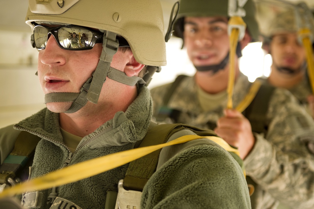 The image shows military personnel in full combat gear, including helmets and tactical equipment, appearing to be inside an aircraft preparing for a parachute jump. The foreground soldier wears reflective sunglasses and a tan helmet with chin strap, holding a yellow static line. At least two additional soldiers are visible in the background in similar gear.
