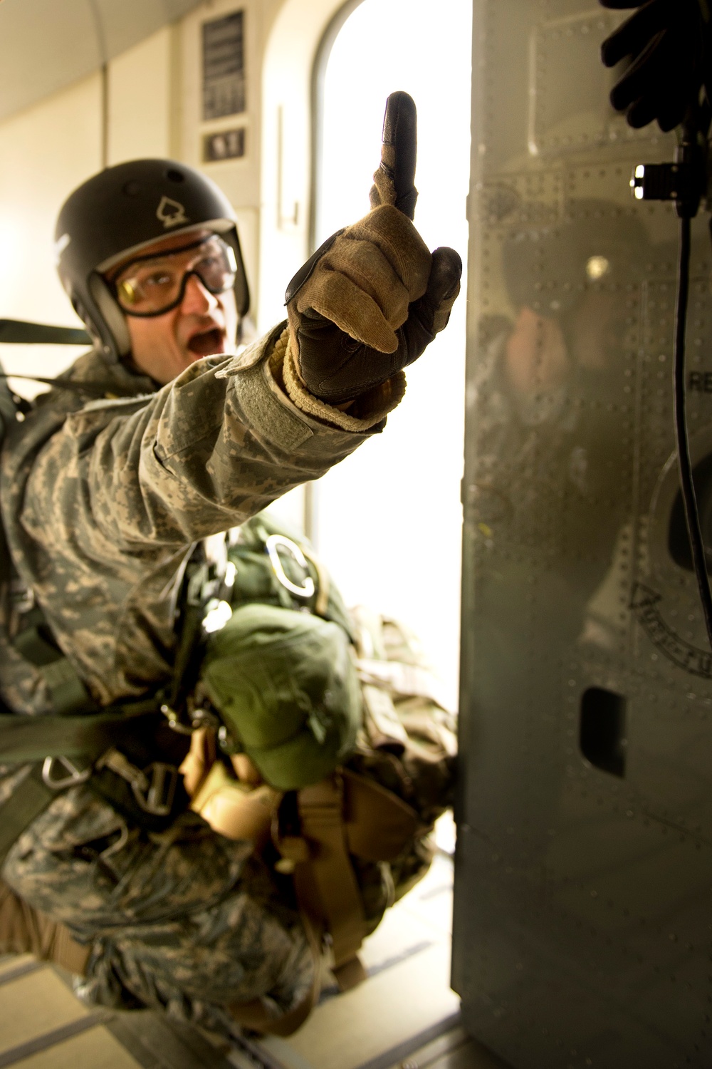A military paratrooper in full jump gear is positioned near an aircraft door, holding up one finger in what appears to be a jump command or countdown signal. The individual is wearing US military digital camouflage (ACU pattern), a black jump helmet with a spade symbol, yellow-tinted protective goggles, and tactical gloves. The setting appears to be the interior of a military transport aircraft during an airborne operation.