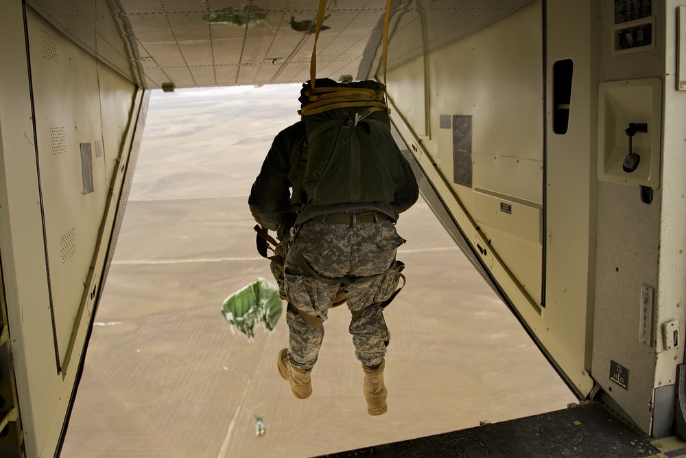 A military paratrooper is captured mid-exit from the open rear ramp of a military transport aircraft over an arid desert landscape. The soldier is wearing US Army combat uniform (ACU pattern) and is equipped with a full parachute rig, with a static line visible connecting to the aircraft's anchor line cable. A pilot chute or deployment bag is visible below and to the left, indicating the parachute deployment sequence has begun.