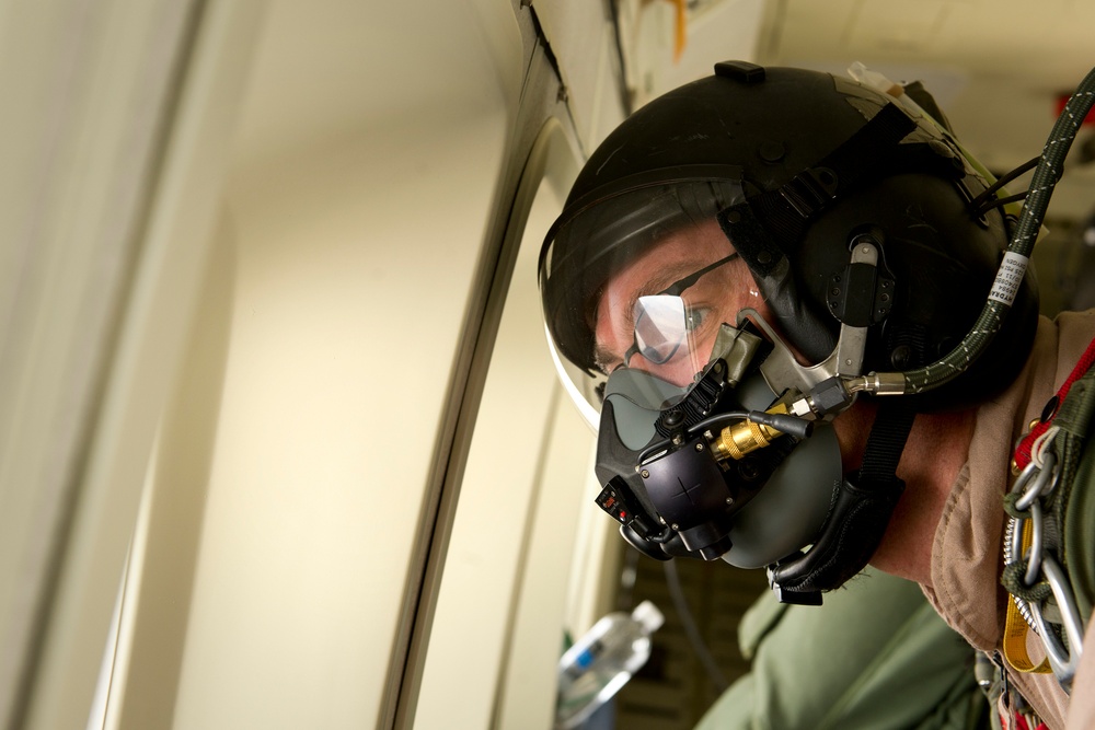 A military aviator or aircrew member is wearing full flight gear including a helmet, oxygen mask, and visor inside what appears to be a military aircraft. The individual is looking to the side, possibly through a window or hatch. The equipment visible is consistent with high-altitude or pressurized flight operations.