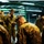 From cold to freezing, Marines head to the Arctic Circle A group of military personnel in digital camouflage uniforms are gathered inside a large industrial or warehouse-type facility with fluorescent lighting. One central figure appears to be reading from a small book or card while others around him have their heads bowed, suggesting a briefing, prayer, or roll call activity. The setting and uniforms are consistent with U.S. Marine Corps MARPAT (Marine Pattern) digital camouflage.