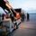 From cold to freezing, Marines head to the Arctic Circle Military personnel in camouflage uniforms are gathered on an airport tarmac around a Delta Airlines baggage conveyor belt vehicle. The scene appears to involve loading or unloading operations, with a cargo container truck visible in the background alongside U-Haul trucks. The photo is taken at dusk or dawn, giving the scene a low-light appearance.