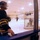 A hockey player in a black and gold jersey and cage helmet stands at the boards watching practice through the glass of an indoor ice rink. On the ice, a goalie guards the net while at least two other players are visible in the background. The image is taken with a wide-angle or fisheye lens, giving it a curved, distorted perspective.