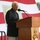 An elderly man with white/gray hair is speaking at a wooden podium aboard the USS Peleliu (LHA), a U.S. Navy amphibious assault ship. He is dressed in a dark blazer and khaki pants, standing before a large American flag backdrop. A map or chart is visible on a board behind him to the left.