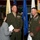 A formal military ceremony featuring two U.S. Marine Corps officers in dress uniforms conversing with a civilian woman holding papers. Multiple military branch flags are displayed in the background, and a decorated cake is visible to the right, suggesting a celebratory event. The setting appears to be an indoor military venue or hangar.