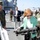 A civilian woman in a green cardigan is handling a military weapon system, likely a rocket launcher or recoilless rifle, during what appears to be a guided tour or demonstration aboard a naval vessel. A U.S. Marine in digital camouflage uniform stands nearby, along with other civilians and military personnel observing the demonstration. The setting appears to be the flight deck of a U.S. Navy amphibious ship.