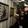 A U.S. Navy officer in dress blues is gesturing toward a framed display board mounted on a ship's interior wall, appearing to brief or explain its contents to an elderly civilian wearing a veteran's cap. The display board contains historical photographs, text, and what appears to be an aerial map, suggesting a commemorative or historical exhibit aboard a naval vessel. The setting appears to be a corridor or passageway inside a warship, likely during a memorial or ceremonial event.